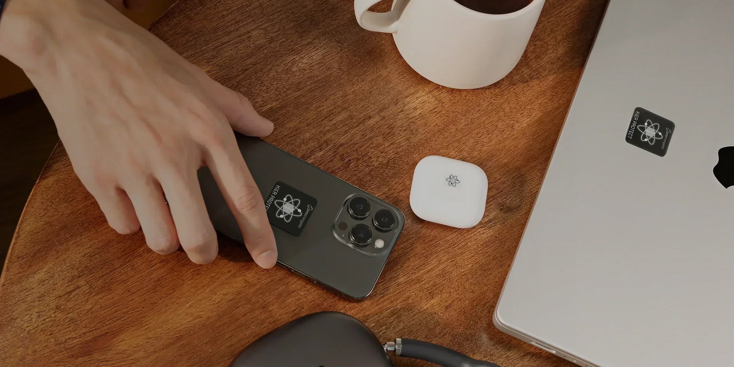 Hand auf Smartphone mit sichtbaren EMF Schutzaufklebern auf Holztisch neben Laptop und Kopfhörern mit EMF Schutzaufklebern sowie Kaffeetasse