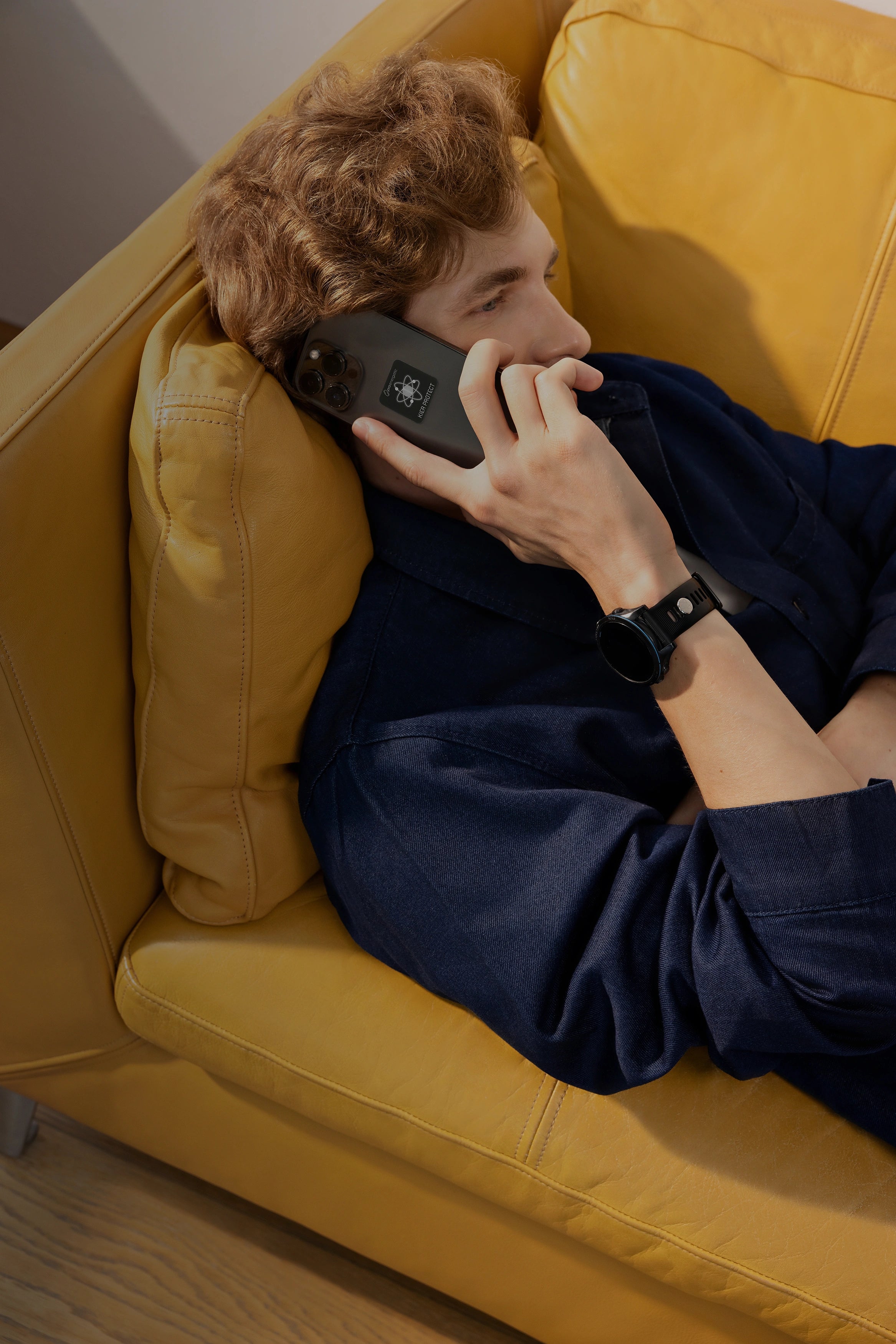 Mann liegt im Innenraum auf einem gelben Sofa, telefoniert mit einem Smartphone mit sichtbaren KIER Protect Mobile Sticker und trägt eine schwarze Smartwatch mit EMF-Protection Pin am Handgelenk