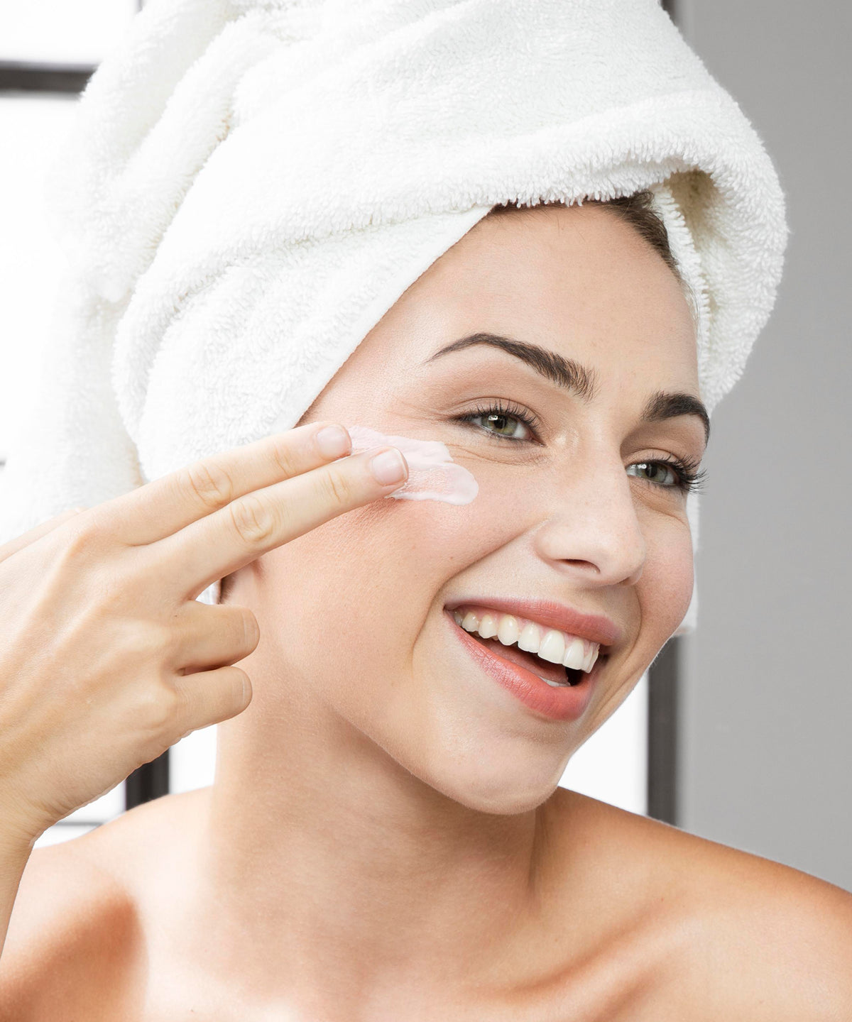 Frau mit Handtuchturban lächelt und trägt Ageless Effect Cream mit den Fingern auf die Wange auf, natürliche Beauty-Anwendung im hellen Badezimmer.
