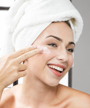 Frau mit Handtuchturban lächelt und trägt Ageless Effect Cream mit den Fingern auf die Wange auf, natürliche Beauty-Anwendung im hellen Badezimmer.