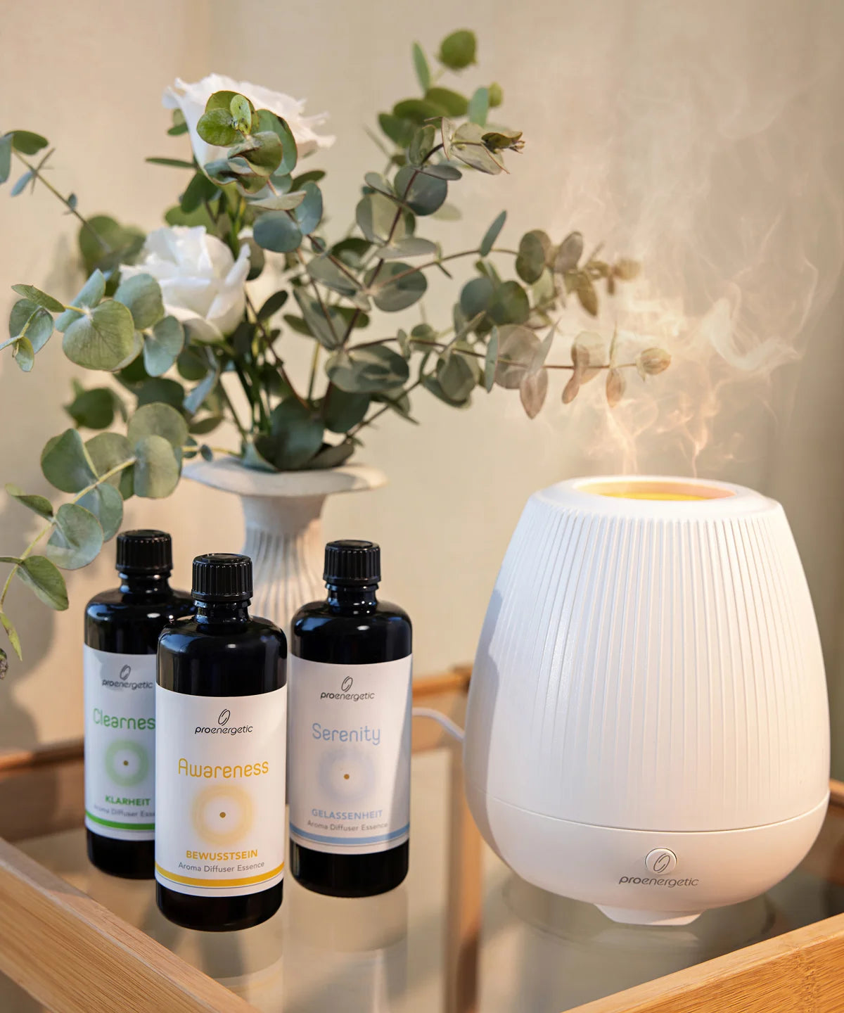 Weißer ProEnergetic Aroma Diffuser auf einem Tisch, daneben drei dunkle Glasflaschen mit ProEnergetic Duftessenzen und eine Vase mit grünen Zweigen im Innenraum