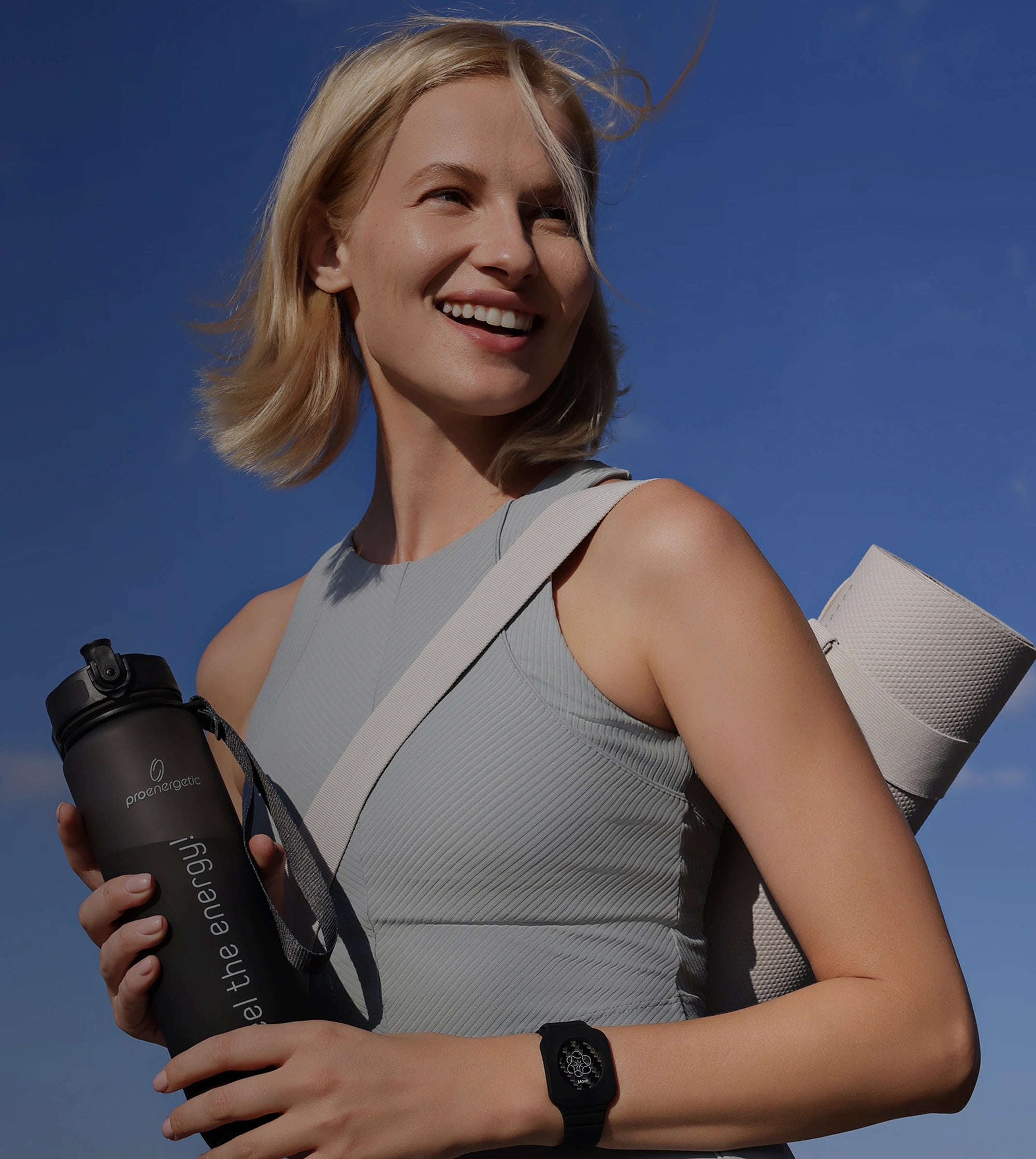 Lächelnde Frau mit umgehängter Yogamatte und schwarzer Pure Move Trinkflasche vor blauem Himmel
