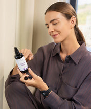 Person sitzt in einem Innenraum und betrachtet eine geöffnete Sprayflasche mit der Aufschrift Clearing Spray in den Händen