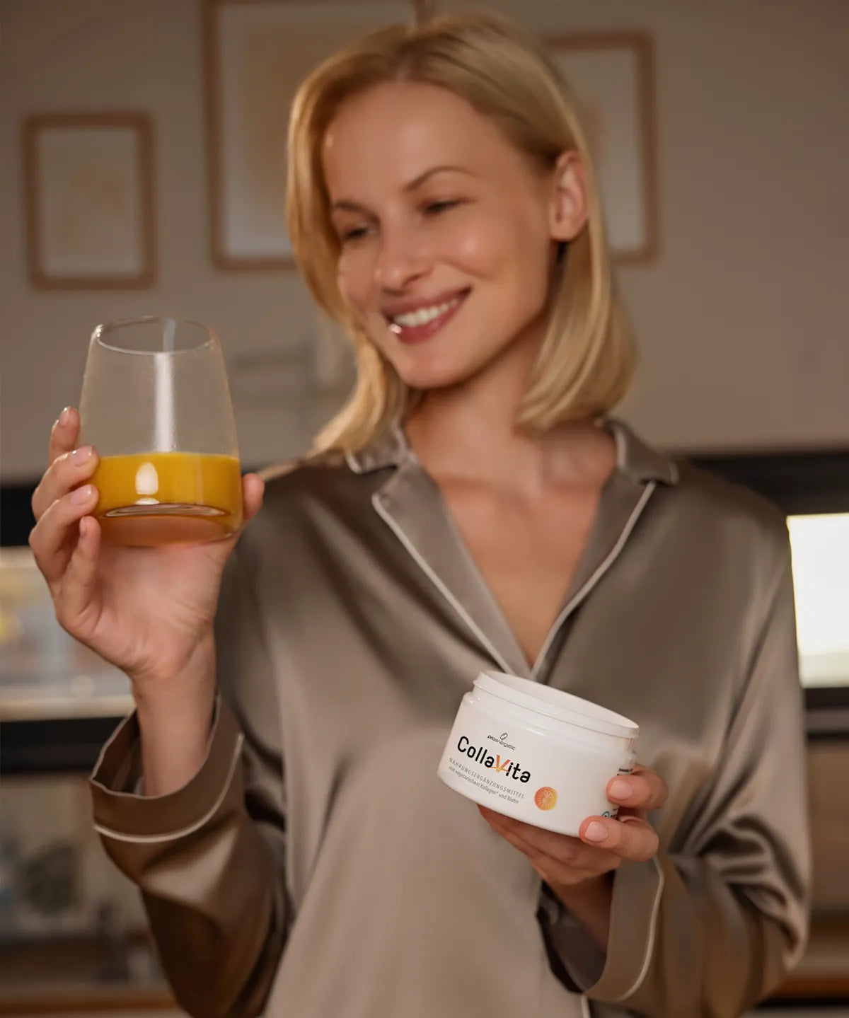 Lächelnde Person in seidiger Loungewear hält ein Glas mit orangem Getränk und eine Dose CollaVita in den Händen, in einer warmen, wohnlichen Küchenumgebung.