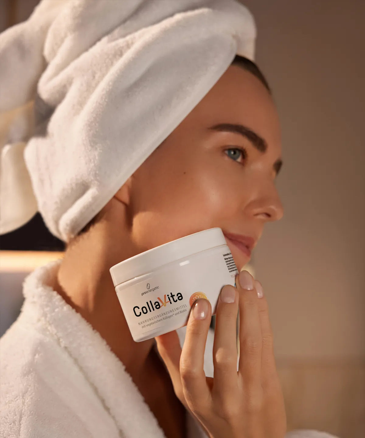 Frau im Bademantel mit Handtuch um den Kopf hält die CollaVita-Dose von ProEnergetic sanft an ihr Gesicht in einer warmen Badezimmer-Atmosphäre.