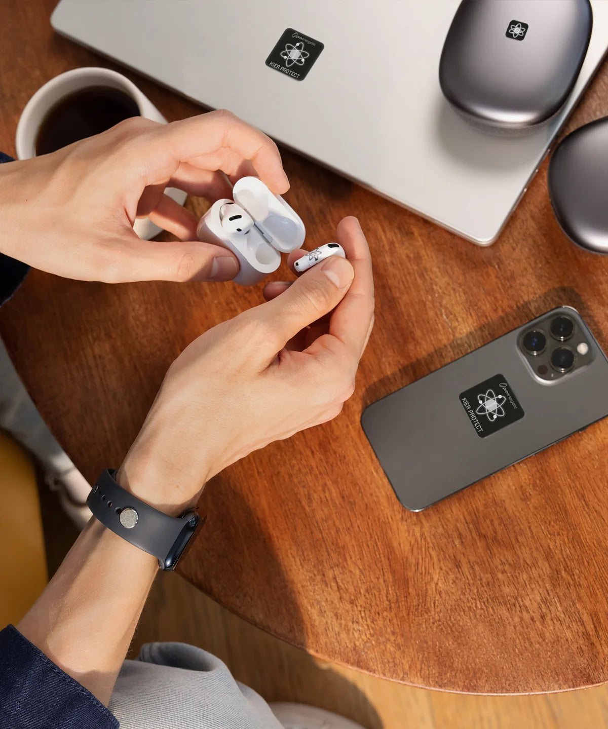 Hände nehmen kabellose In-Ear-Kopfhörer aus einem Ladecase. Auf dem Holztisch liegen Smartphone und Laptop, beide mit E-Protect-Audio-Aufklebern versehen, daneben eine Kaffeetasse.