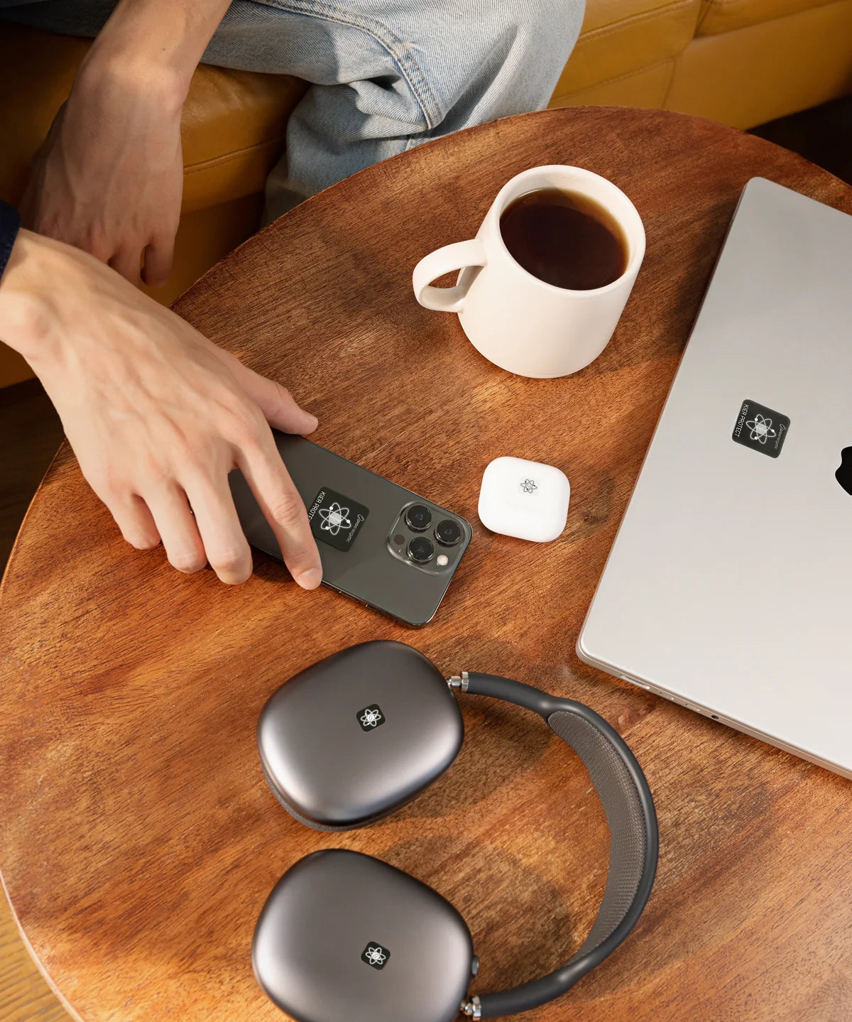 Auf einem runden Holztisch liegen Smartphone, Laptop, kabellose Kopfhörer und ein Ladecase, jeweils mit einem schwarzen E-Protect-Audio-Aufkleber versehen. Daneben stehen eine Tasse Kaffee und eine Hand, die das Smartphone berührt – Alltagsszene im Café- oder Wohnambiente.
