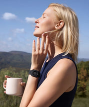 Frau steht im Freien mit einer Tasse in der Hand und trägt ein schwarzes Eloop Band Silikonarmband mit Calm Chip am Handgelenk vor Landschaftshintergrund