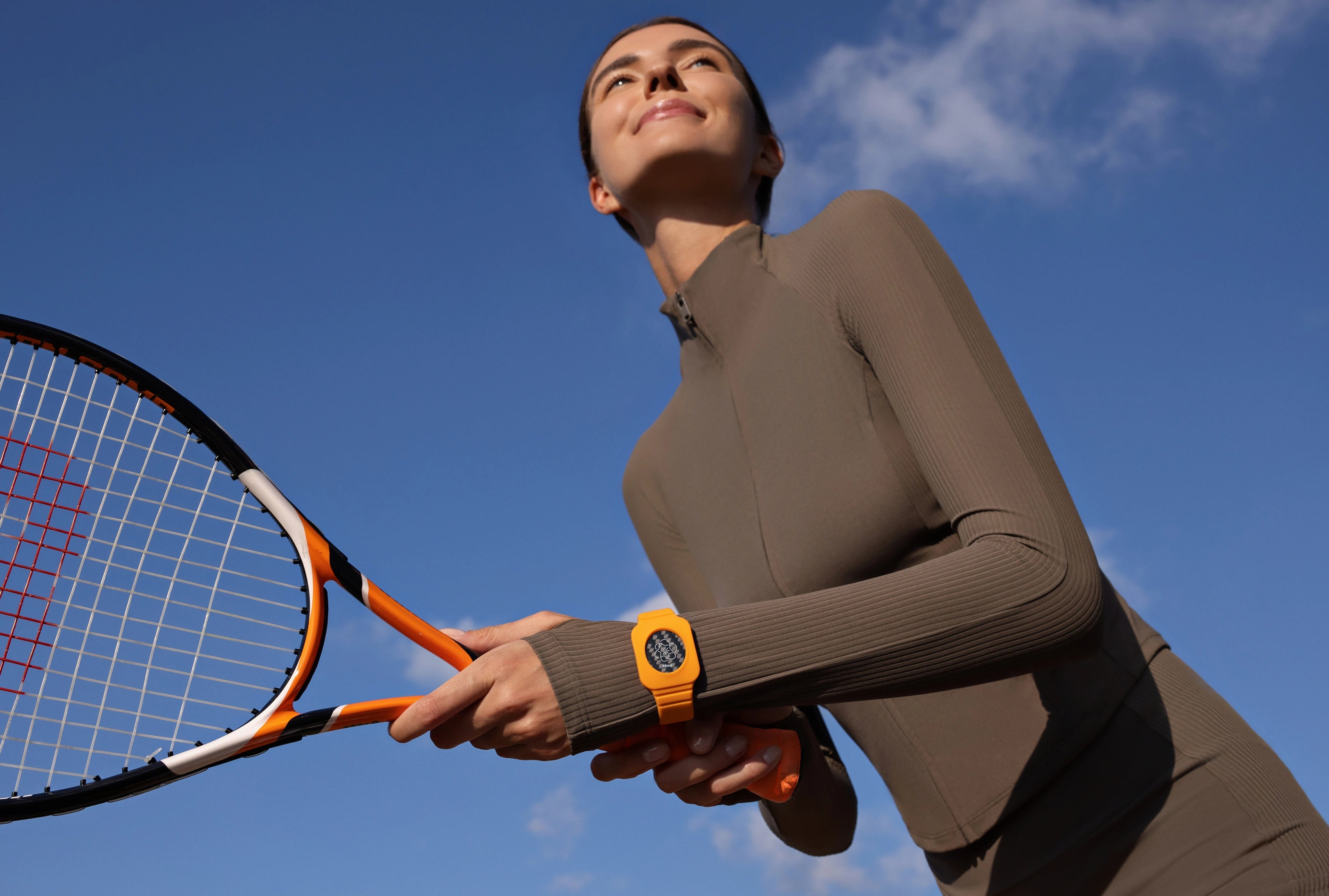 Person hält einen Tennisschläger im Freien und trägt ein oranges Eloop Band Silikonarmband am Handgelenk vor blauem Himmel
