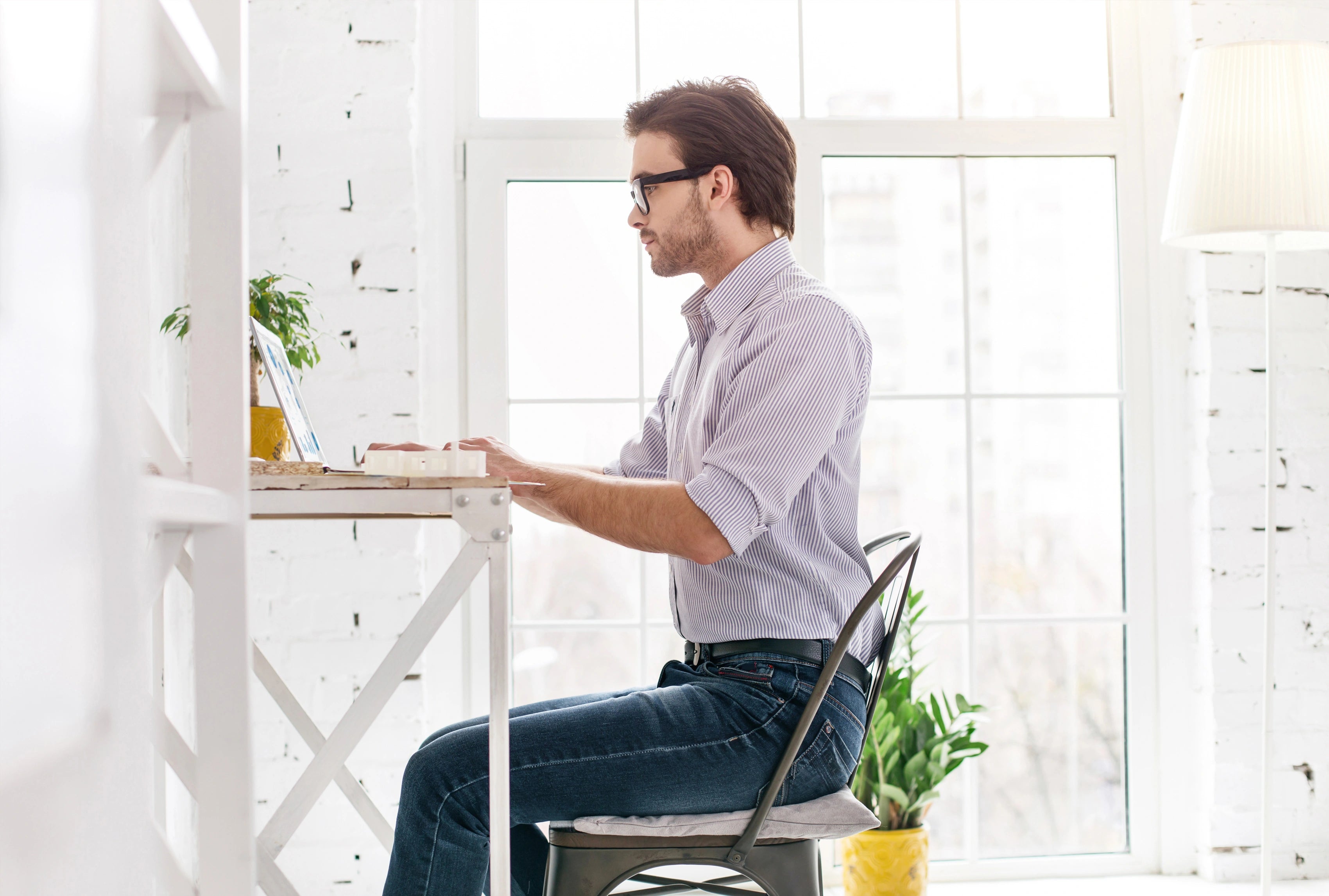 Person sitzt an einem Schreibtisch am Fenster und arbeitet am Laptop, auf dem Stuhl liegt ein weißes Grounding Relax Pad
