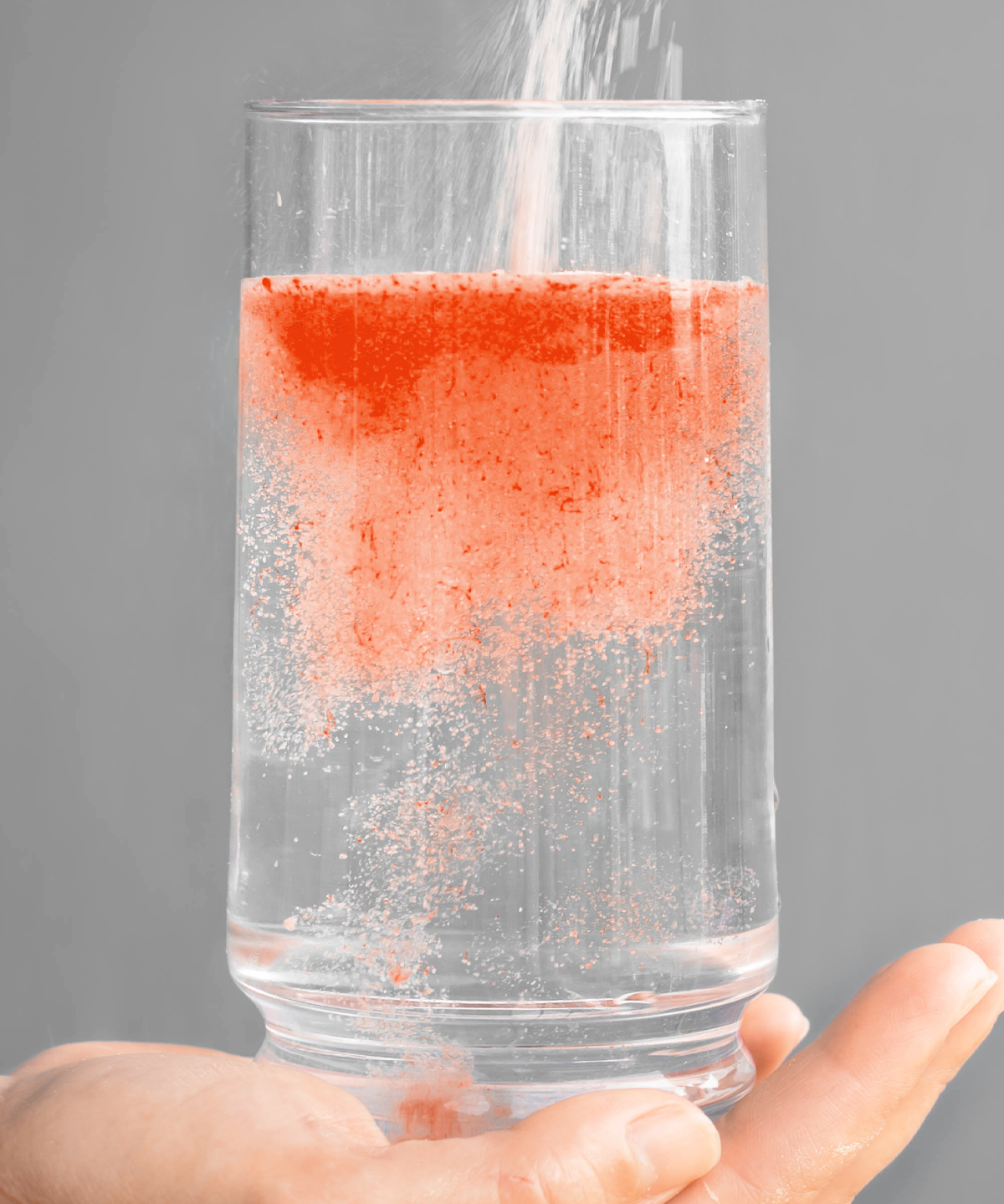 Ein Glas Wasser wird gehalten, während ISOmove Fit-Pulver hineingestreut wird. Das orangefarbene Pulver löst sich sprudelnd auf und färbt das Wasser gleichmäßig – symbolisiert schnelle Löslichkeit, Frische und den Blutorangen-Geschmack.
