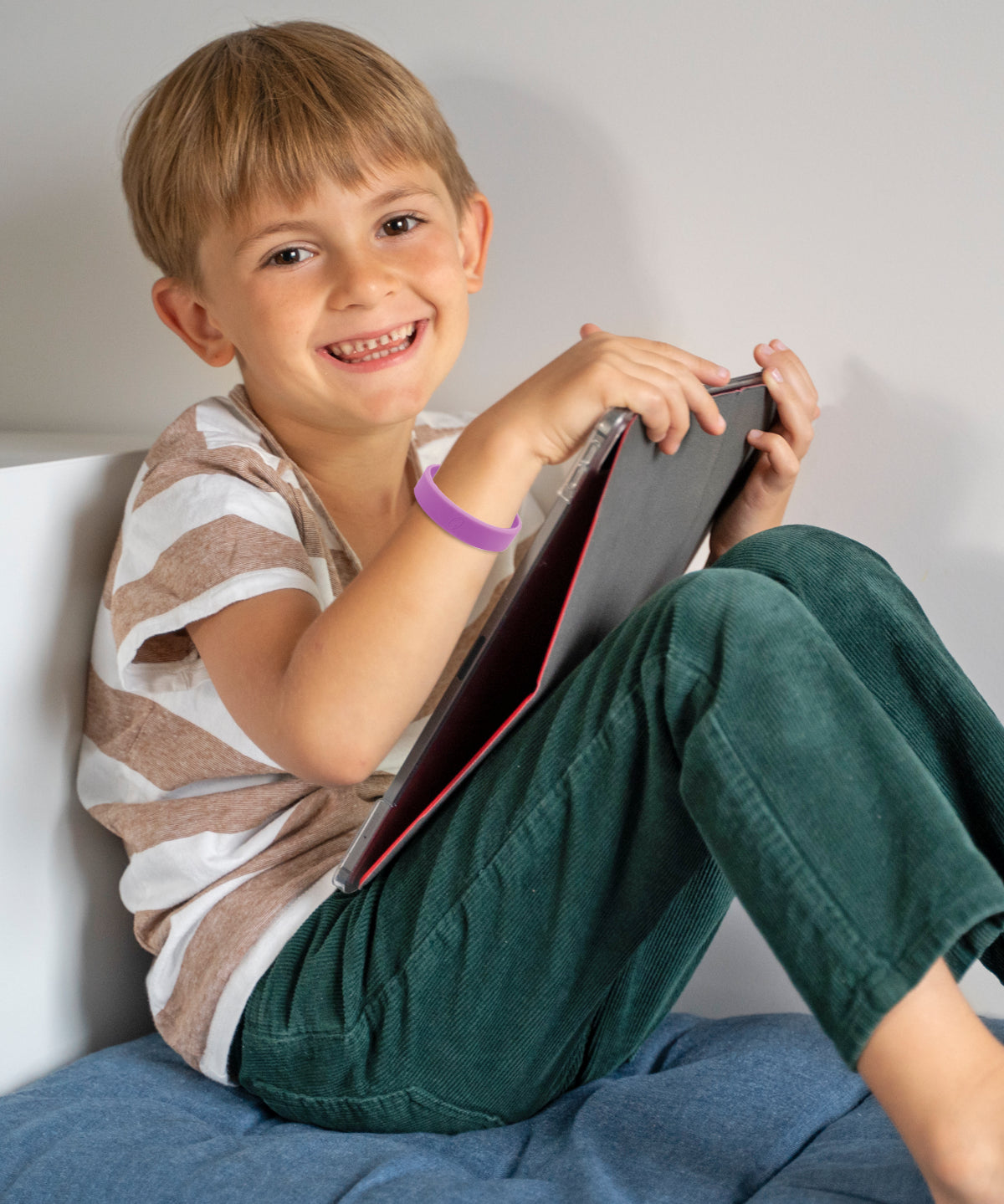 Junge sitzt im Innenraum und hält ein Tablet, am Handgelenk trägt er ein lila ProEnergetic Kids Band aus Silikon