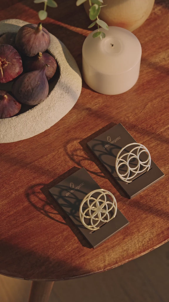 Ein Flower of Love Goodie und ein Seed of Happiness Goodie als goldene Ornamente auf schwarzen Sockeln, arrangiert auf einem Holztisch neben Kerze und Obstschale