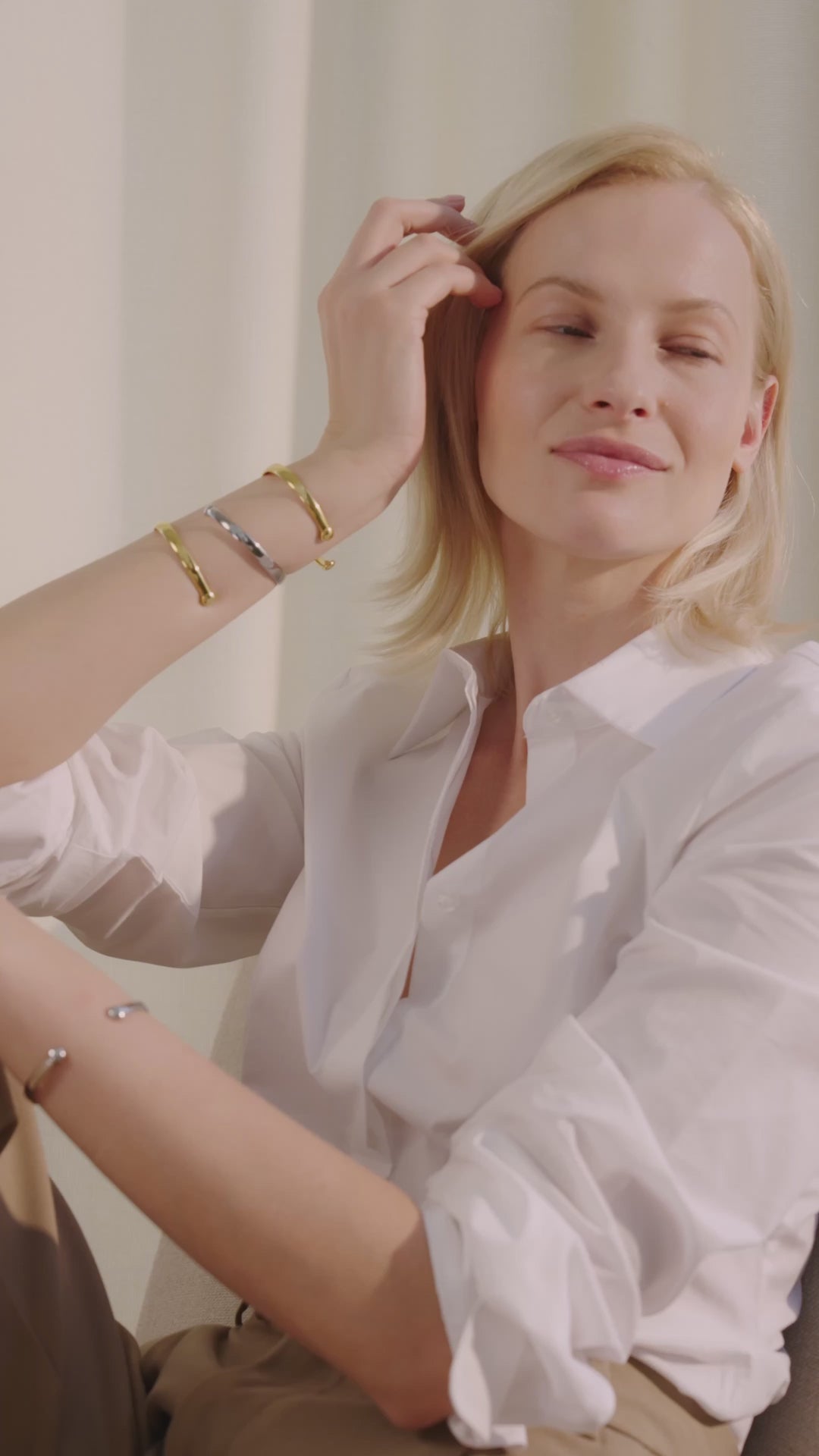 Frau sitzt in einem Innenraum auf einem Stuhl, lächelt und trägt die Lunara Cuff Armreifen in Gold und Silber mehrfach am Unterarm und Handgelenk