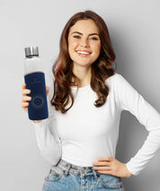 Person steht vor hellem Hintergrund und hält den Water Vitalizer in der Hand, Glastrinkflasche mit dunkelblauer Schutzhülle sichtbar